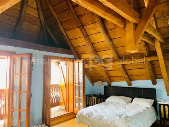 Dormitorio doble con dos ventanas que dan al balcón y vigas de madera y paneles del techo abuhardillado.